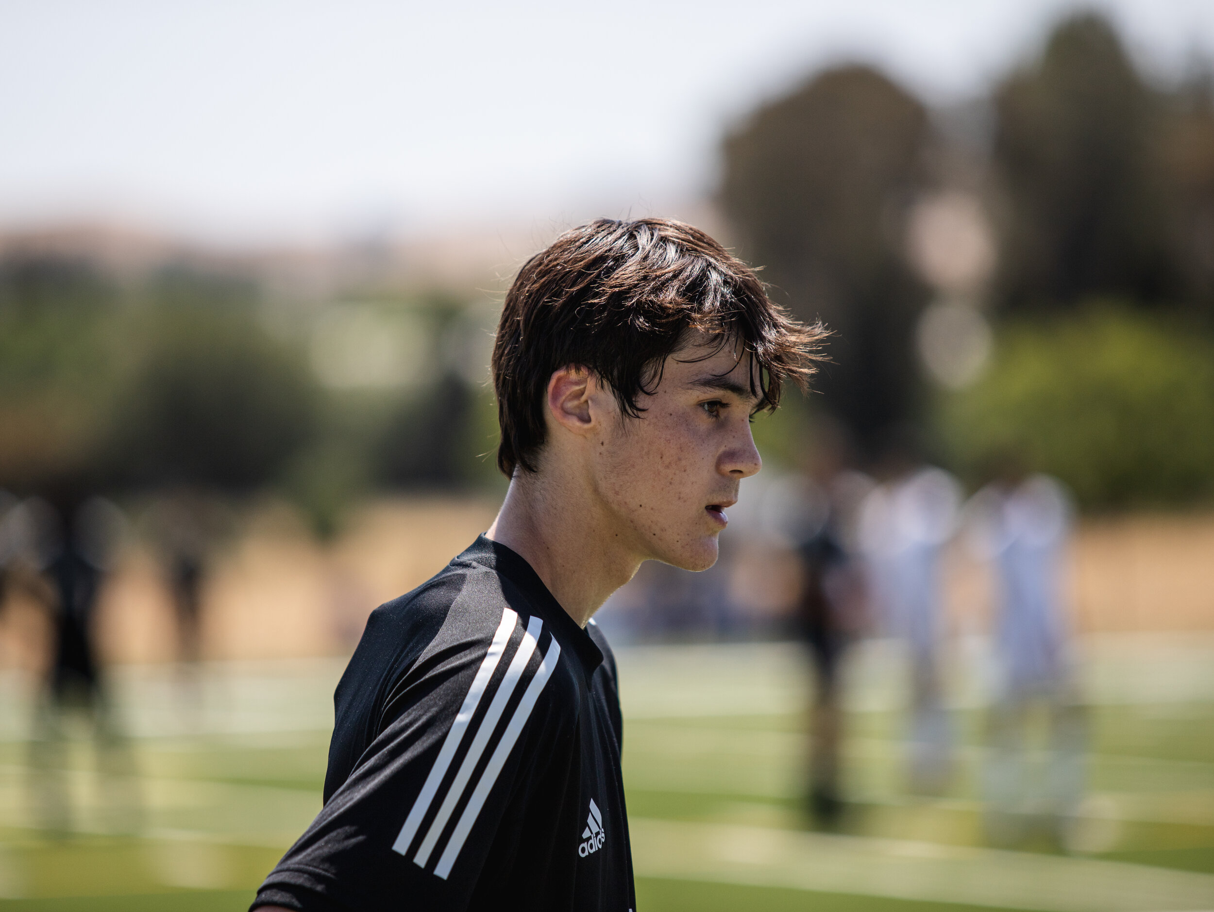 Side profile of a teenage boy with dark brown hair in a black Adidas sports shirt with white stripes, outdoors on a sunny day, with a blurred background of people and trees.
