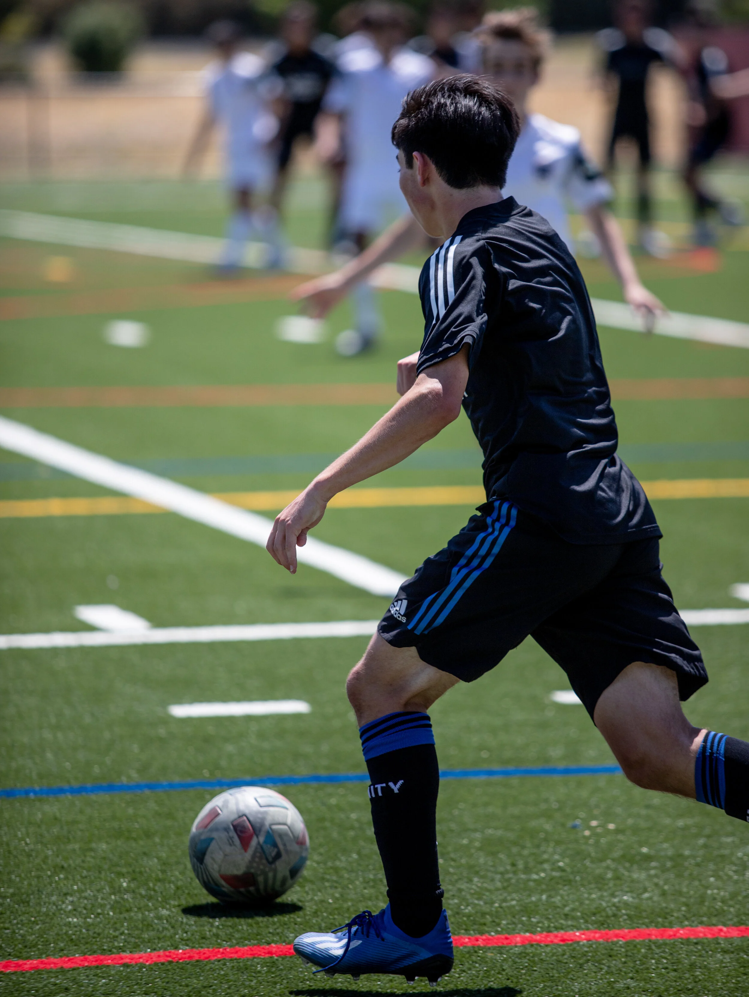 A young male soccer player dressed in black with blue stripes, wearing black knee-high socks and blue cleats, is preparing to kick a soccer ball on a green turf field during a game.