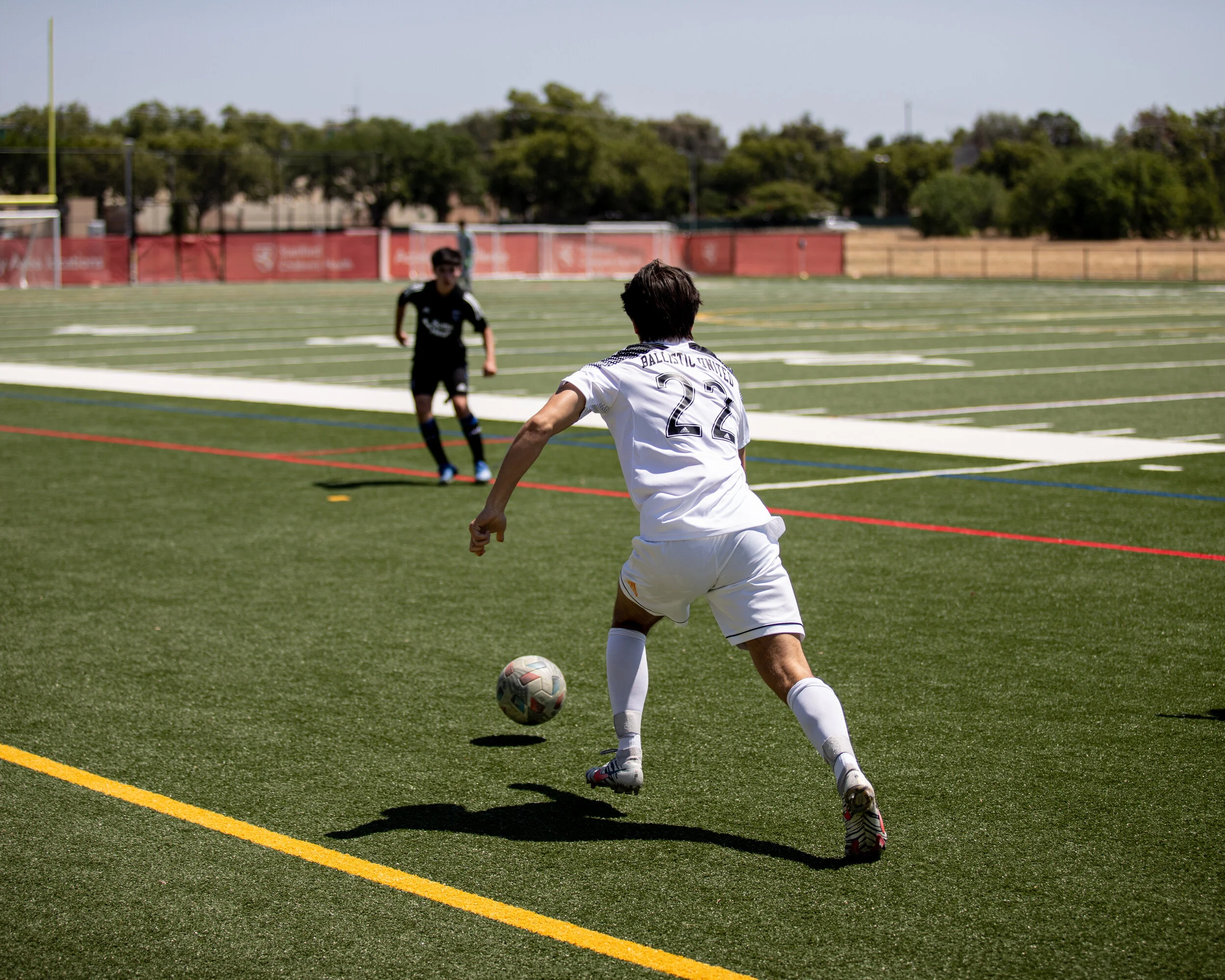 Soccer player in white uniform preparing to kick the ball on a green turf field during a game with another player in black in the background.