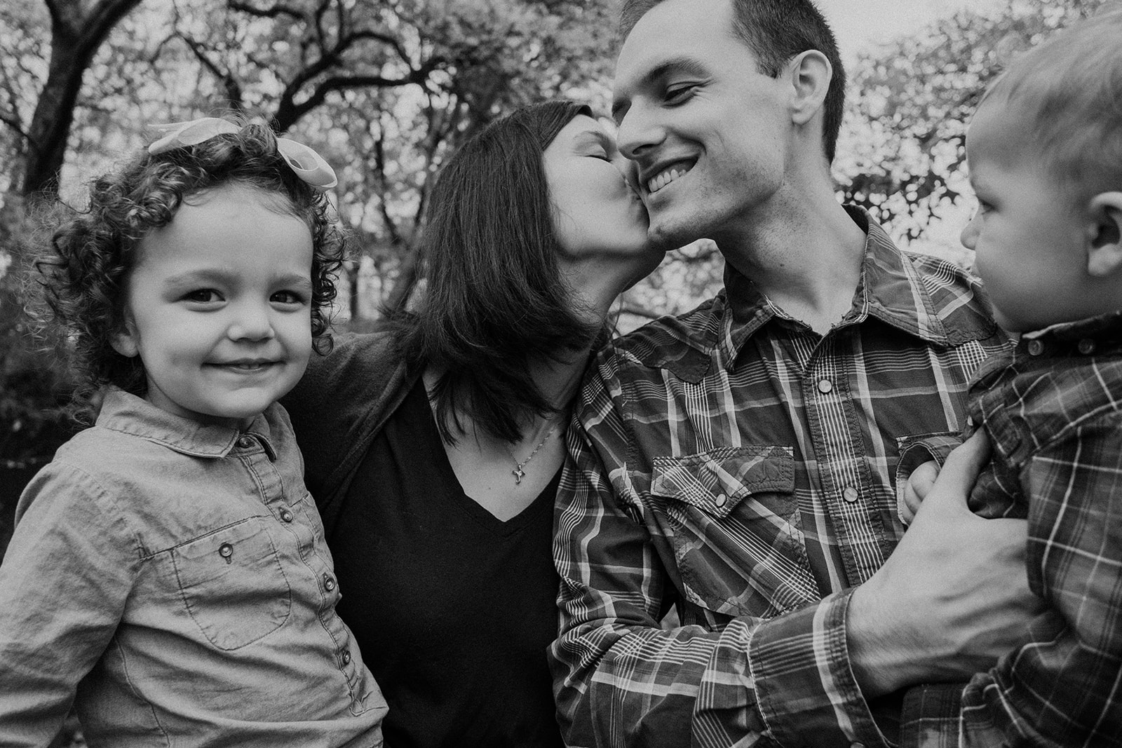 A mother kisses her husbands cheek while her children smile.