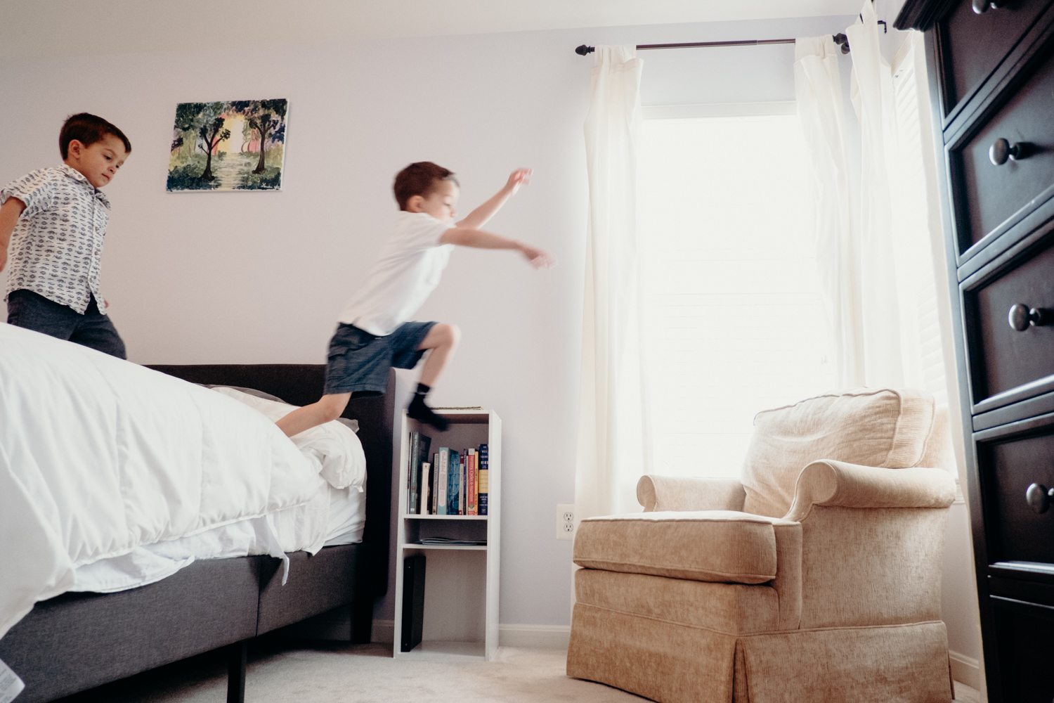 A little boy jumps off his parents bed onto a chair while his little brother watches.
