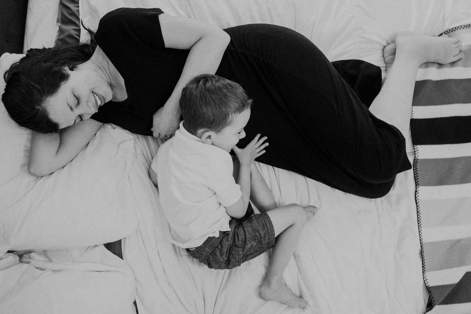 A little boy touches his mother's pregnant belly and listens for baby to make noises while his mother lays in bed.