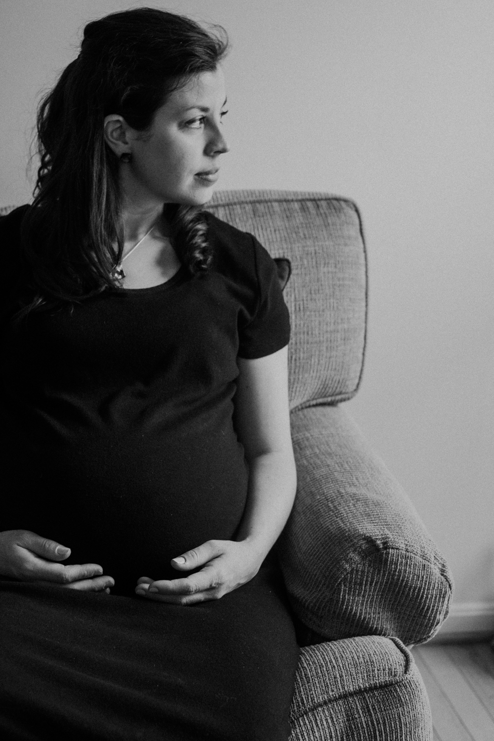 A pregnant mother sits on a couch and looks out the window while holding her belly. 