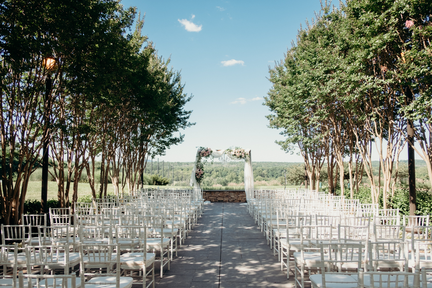 Rows of white chairs with a center aisle leads up to an arbor decorated with while fabric and flowers for a wedding at Lansdowne Resort.