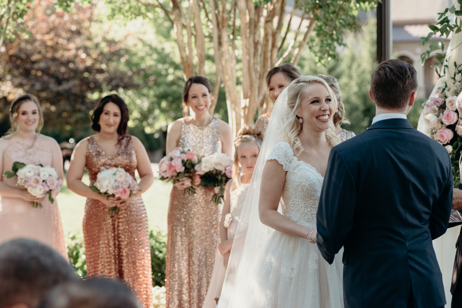 A bride smiles at her groom while her bridesmaids in sparkly dresses look on during a wedding ceremony at Lansdowne Resort. 
