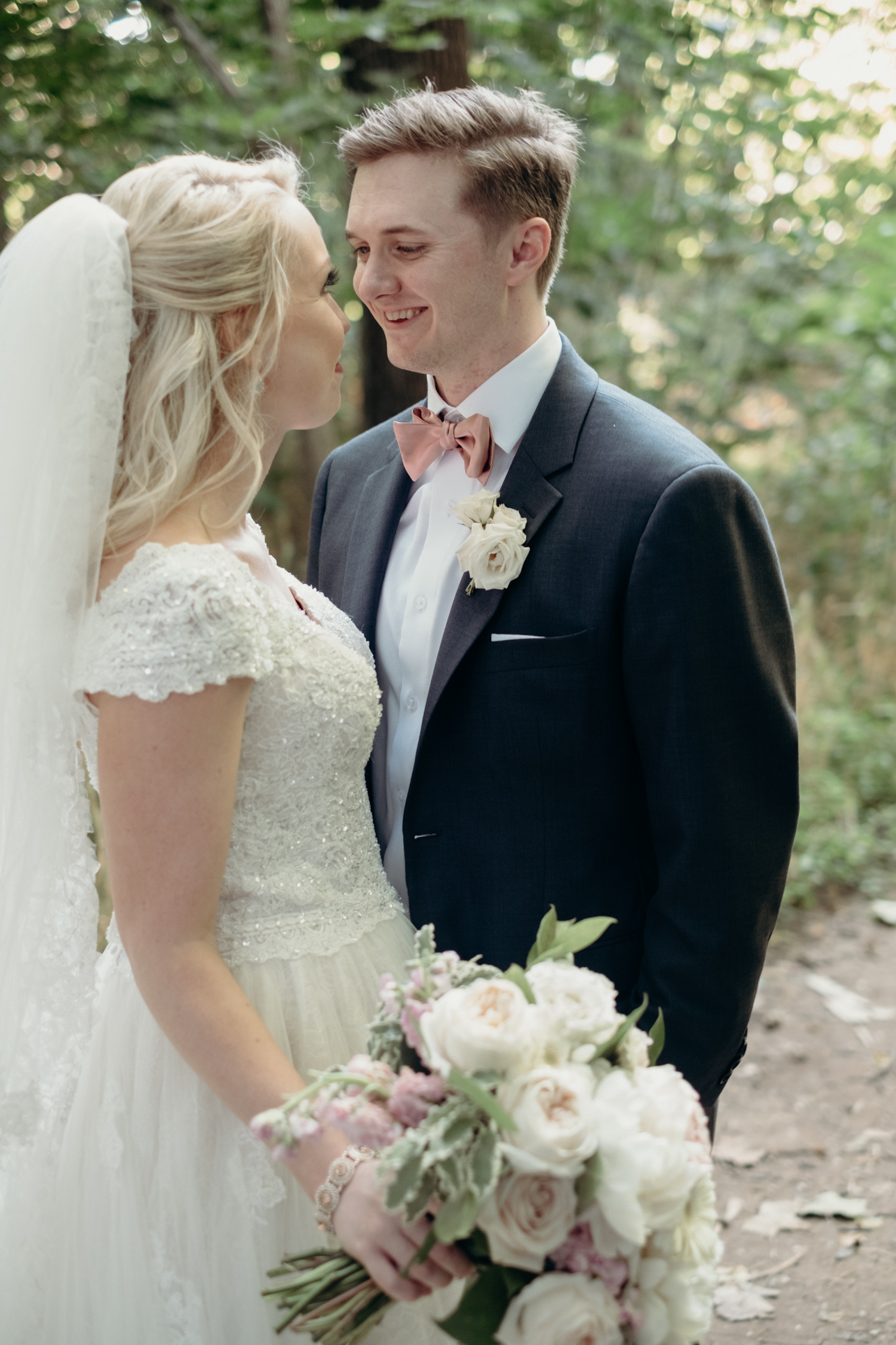 A bride carrying a bouquet of white and pink flowers looks into the eyes of her groom in a peach bowtie in the woods at Lansdowne Resort and Golf Course. 