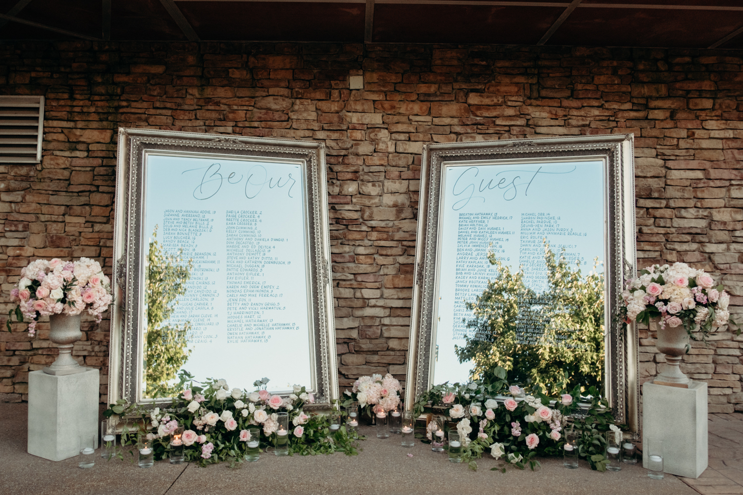 A seating chart for guests at a wedding reception is written on two large mirrors surrounded by lush florals.  