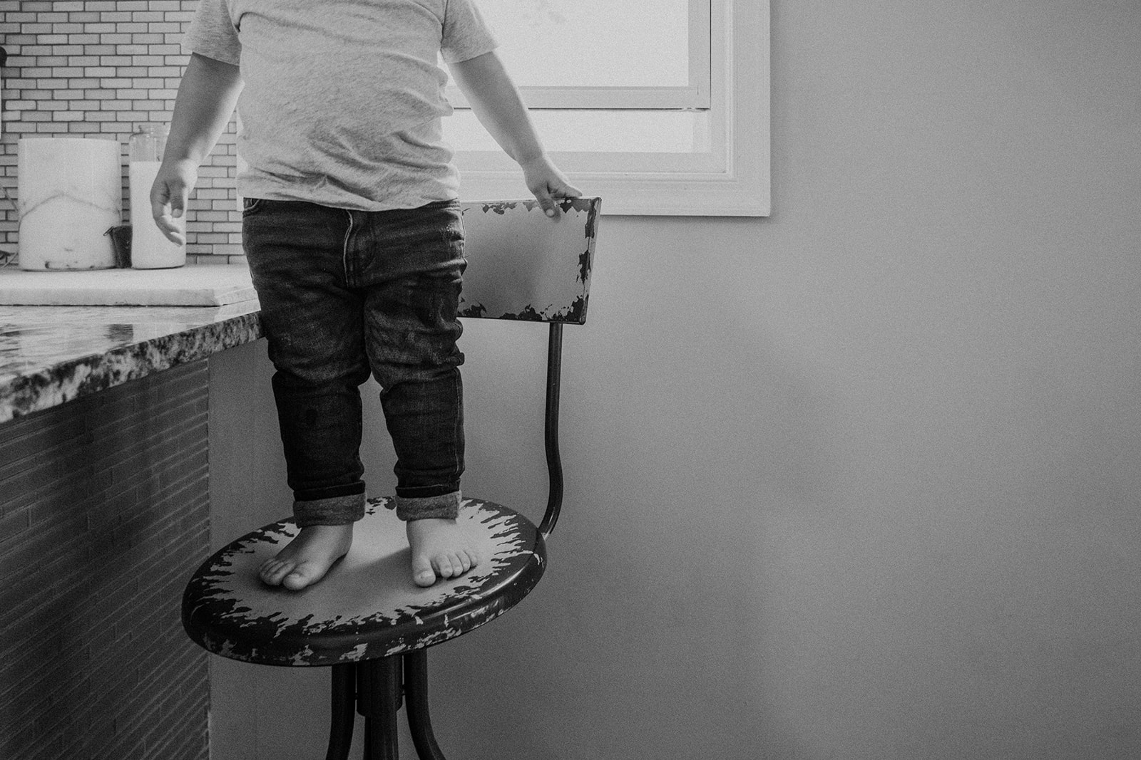A little boy stands on a chair by the kitchen island in his home during an in-home family photography session. 