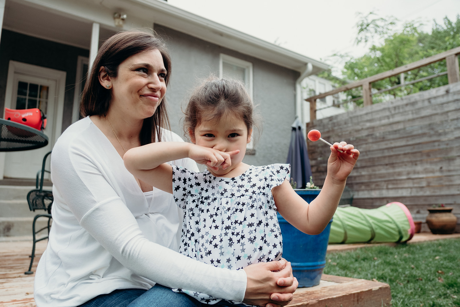 A little girl holds up her lollipop while sitting with her mother on the back deck of their DC home during an in-home family photography session.