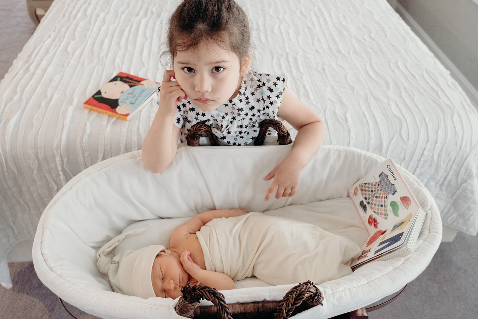 A little girl sits next to her baby brother in his bassinet during an in-home family photography session.