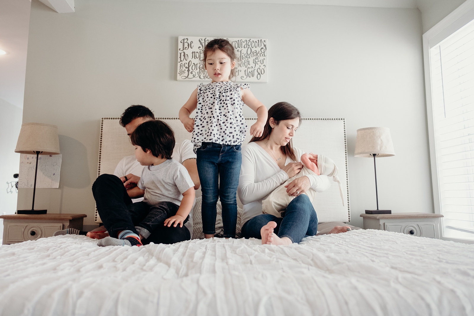 A little girl stands up on her parents bed while the rest of her family sits during an in home family photography session. 