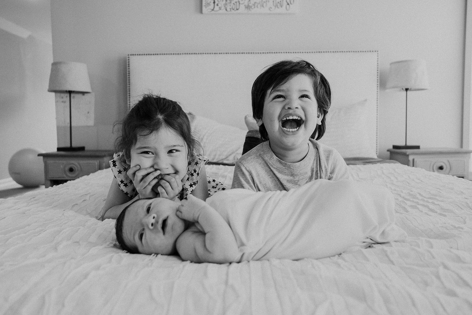 A brother and sister giggle while their baby brother lies in front of them during an in home family photography session. 