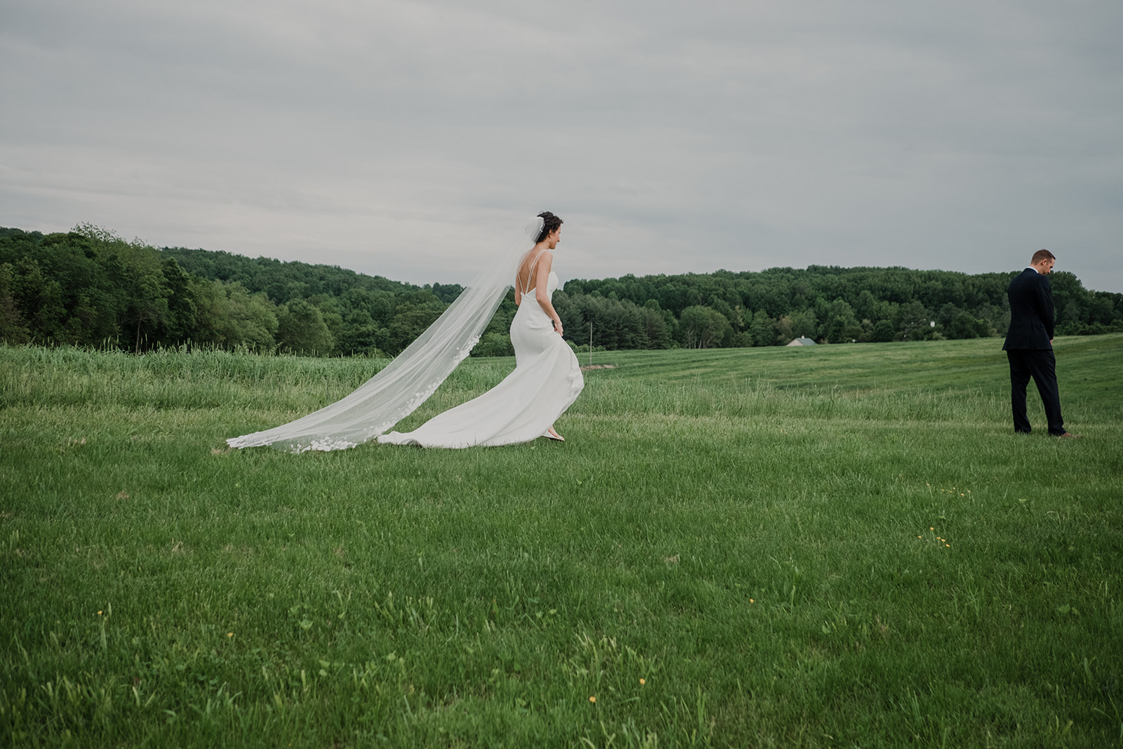 A bride with a long veil walks through a field towards her groom for a first look before their outdoor wedding ceremony at Blue Hill Farm in Waterford, VA.