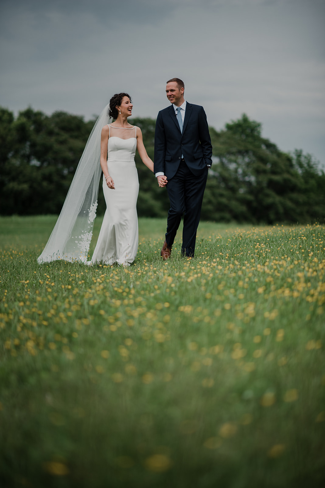 A bride and groom hold hands and walk through a field with buttercups before their outdoor wedding ceremony at Blue Hill Farm in Waterford, VA. 