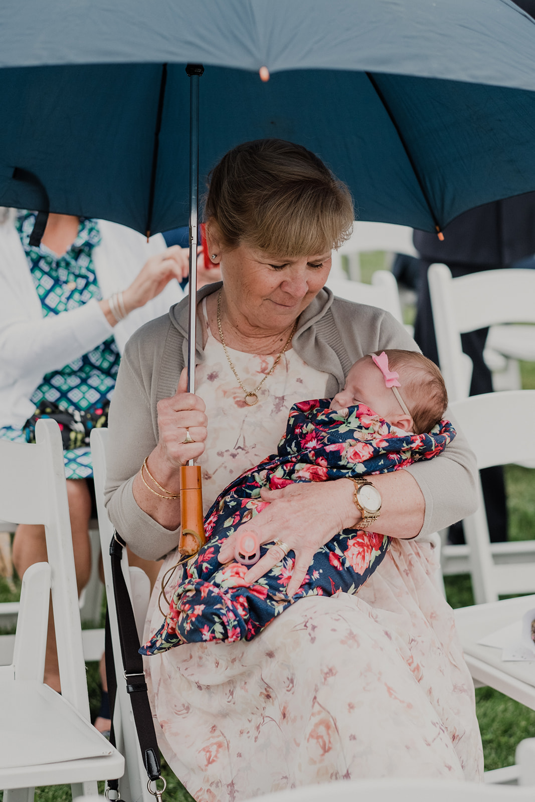 A grandmother holds her swaddled granddaughter under an umbrella at an outdoor wedding ceremony at Blue Hill Farm in Waterford, VA. 