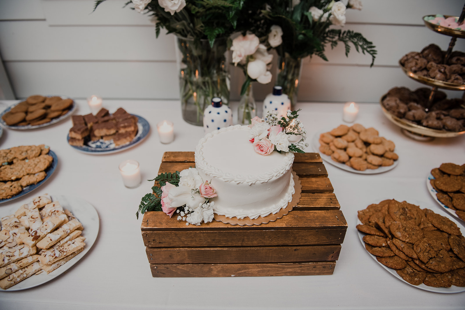 A white buttercream cake sits on a rustic wooden crate surrounded by other desserts at a wedding reception at Blue Hill Farm in Waterford, VA.