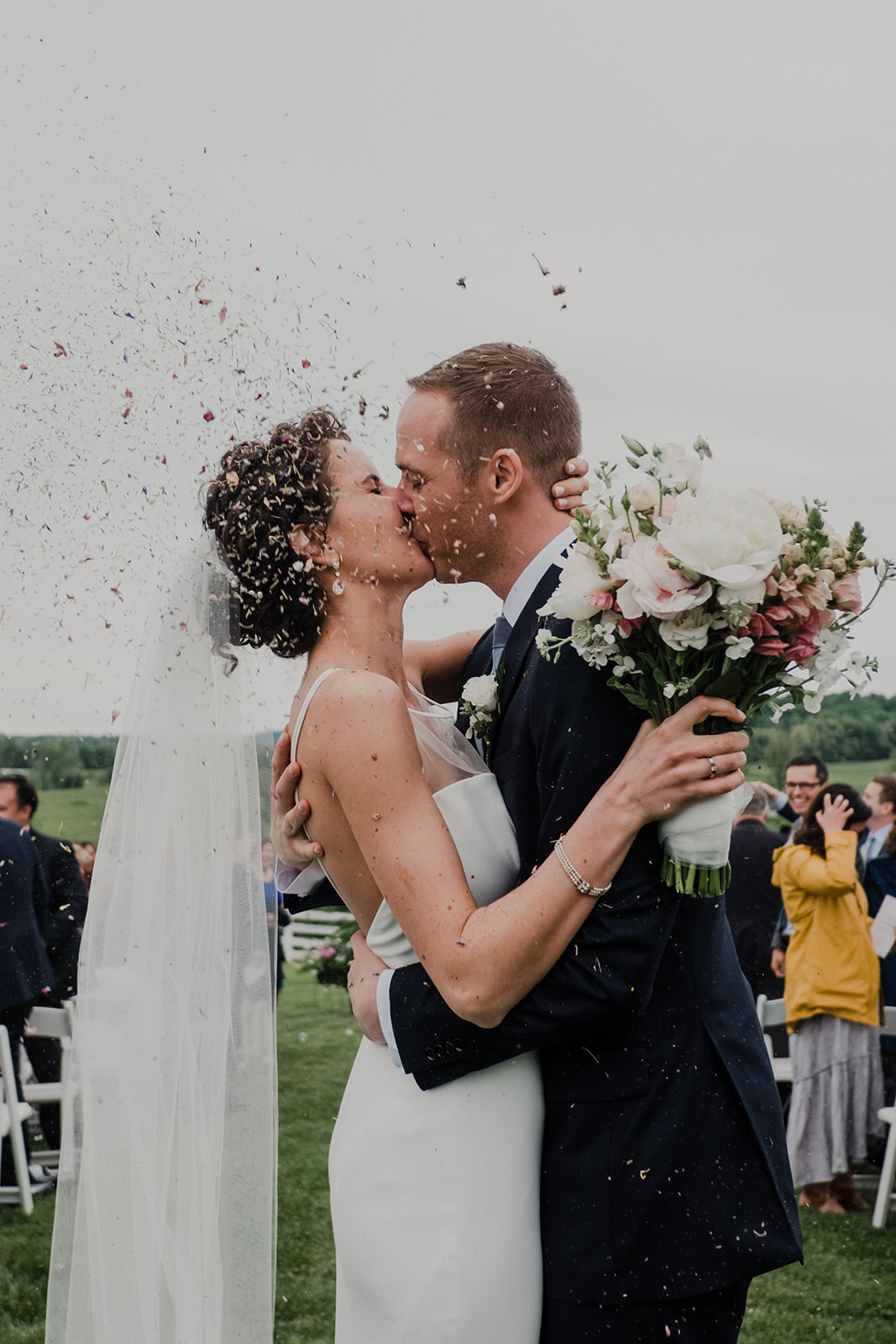 A bride and groom kiss in the aisle after their outdoor wedding ceremony at Blue Hill Farm in Waterford, VA as guests toss flower petal confetti.  