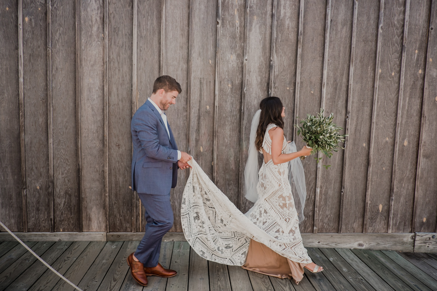 groom holds dress for bride as they walk across the docks after first look