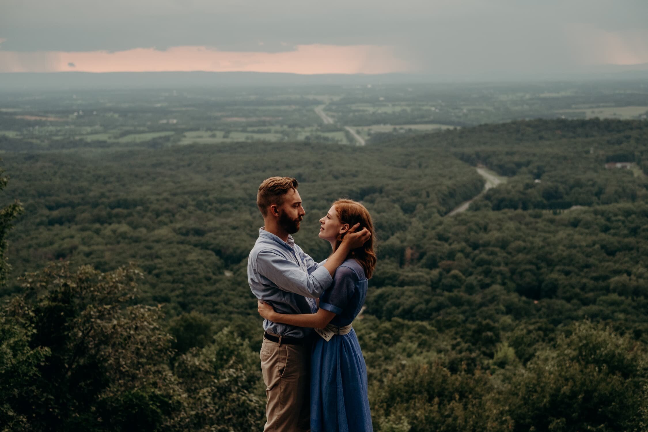 Couple holds each other on cliff with thunderstorms at sunset in background