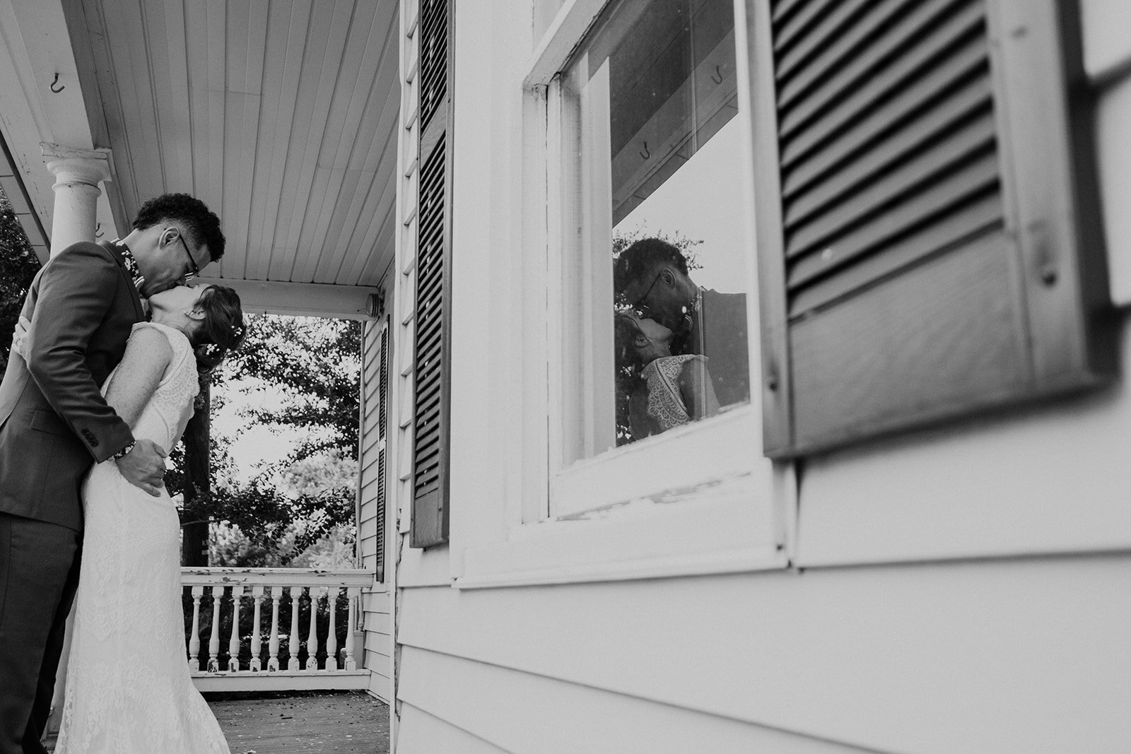 A bride and groom's image is reflected in a porch window while they kiss. 