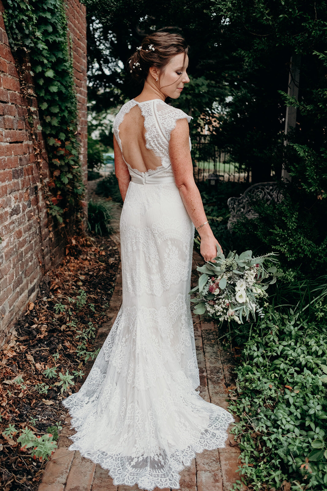 A bride with a sleeveless, open back lace wedding dress looks down at her bouquet.