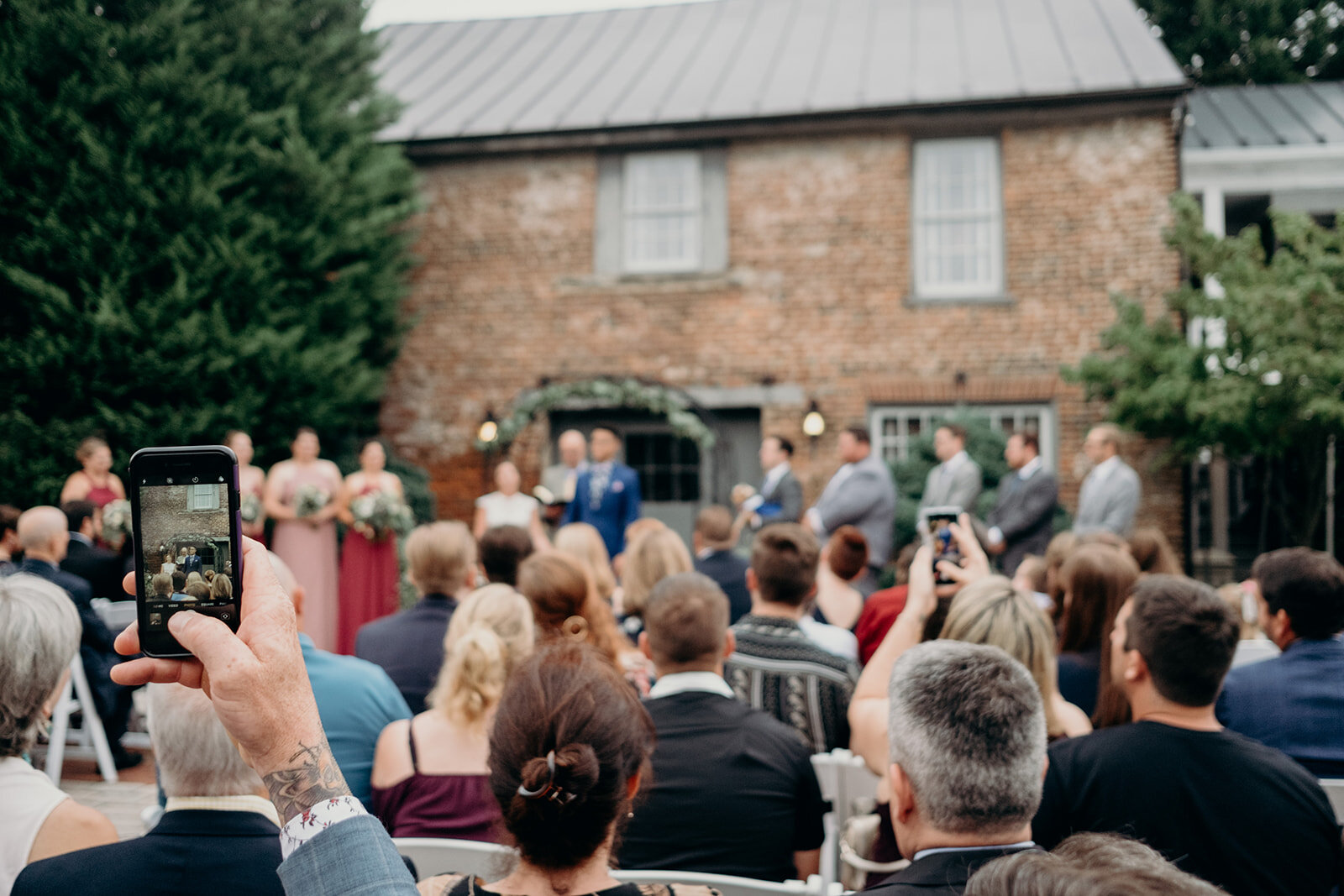 A tattooed guest takes a phone photo of the bride and groom after they are declared husband and wife during their outdoor wedding ceremony at Birkby House in Leesburg, VA. 