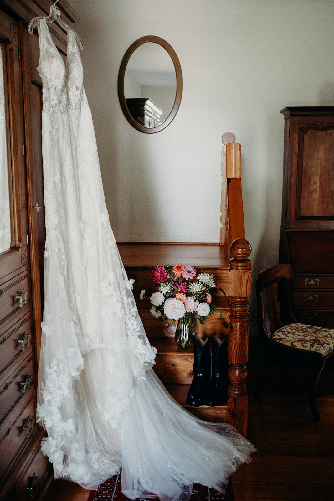 A long lace wedding dress hangs next a bright bouquet and the groom's black cowboy boots before an outdoor farm wedding. 