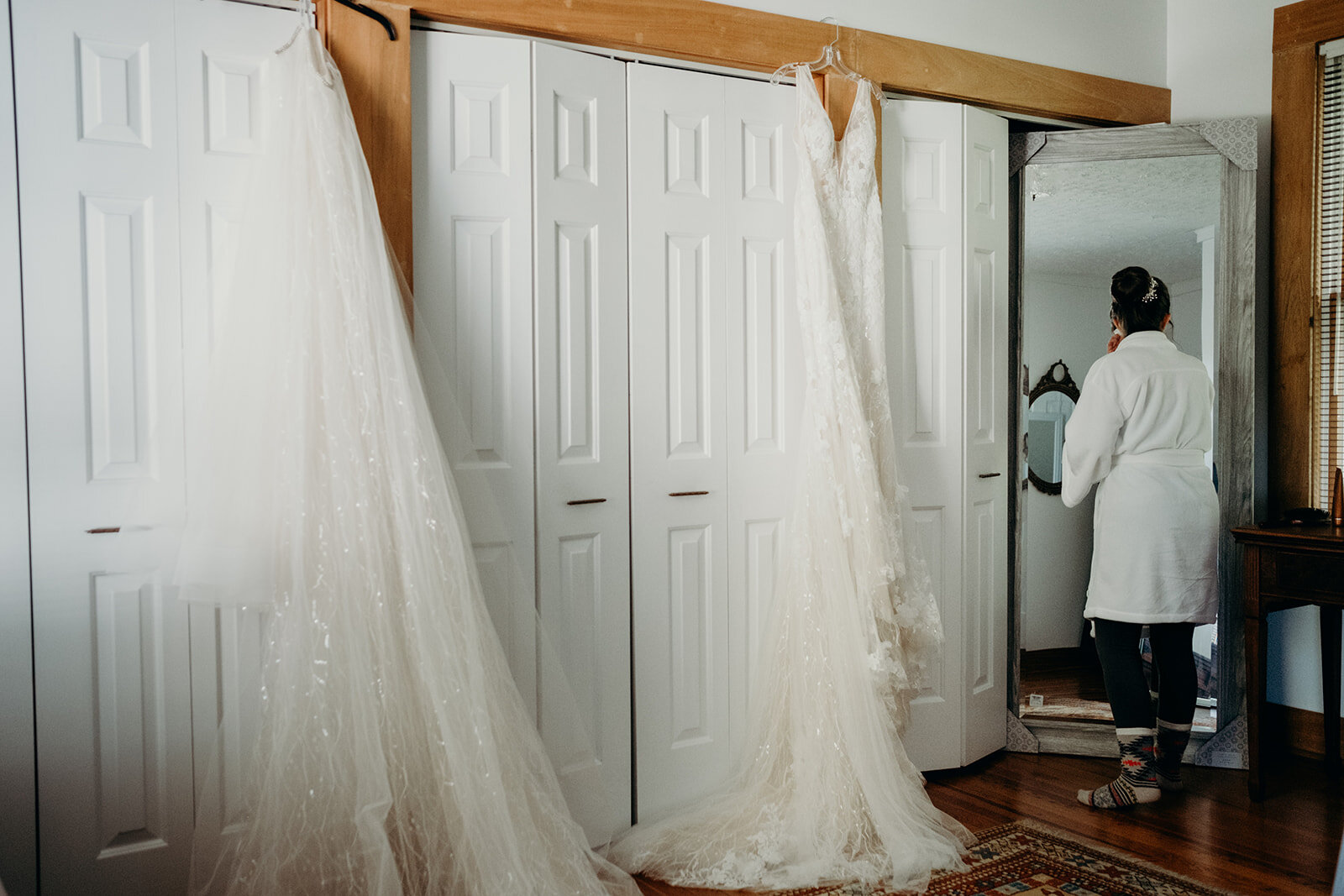 A bride peeks in the mirror while her wedding dress and overskirt hang nearby.