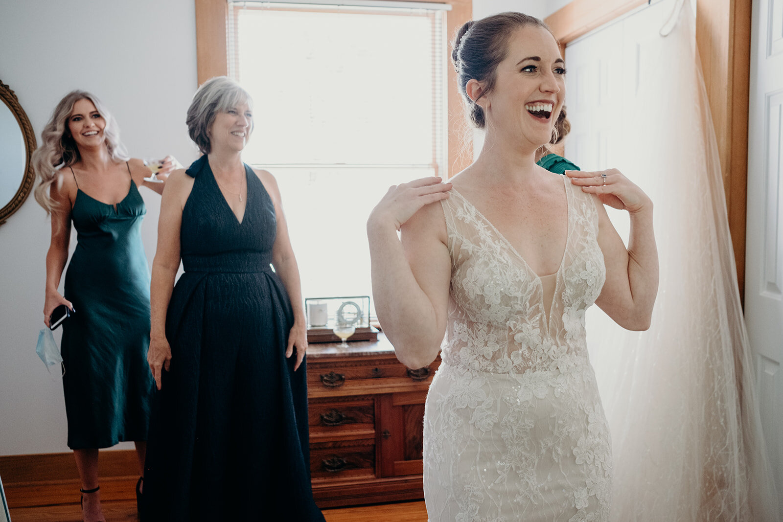 A bride smiles after putting on her wedding dress while her mother and sister look on. 