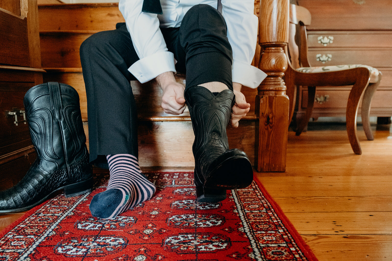 The groom puts on his black cowboy boots in an old farmhouse before his outdoor wedding ceremony.
