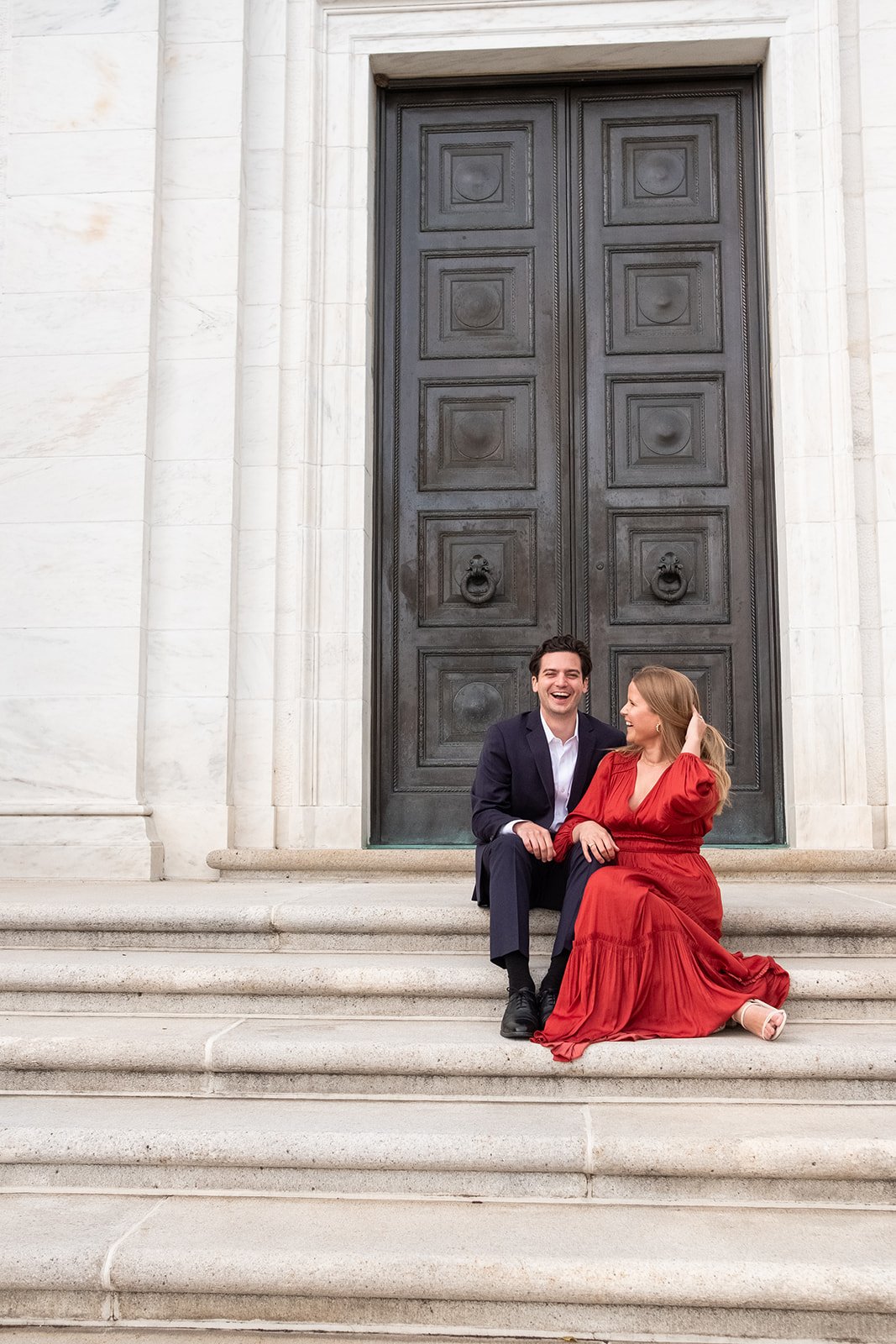An engaged couple sits in front of the bronze doors at the American Institute of Pharmacy Building in Washington, DC.