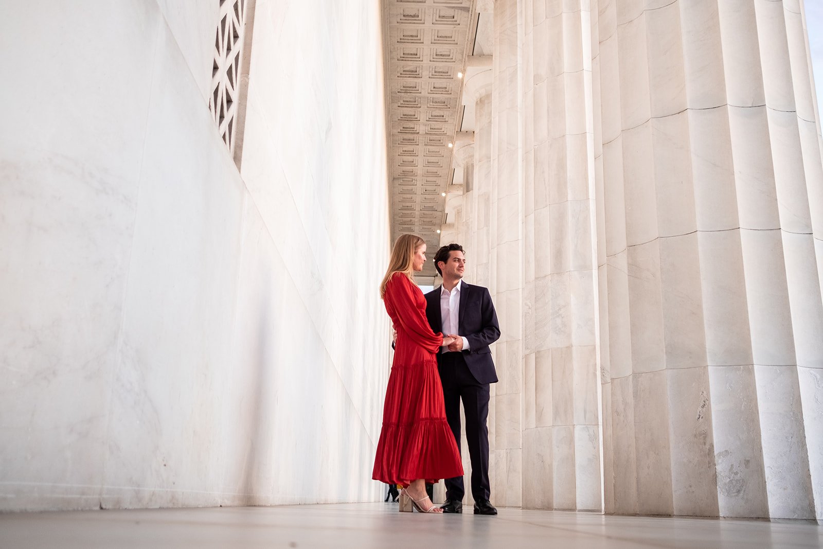 An engaged couple holds onto one another by the columns of the Lincoln Memorial in Washington, DC.