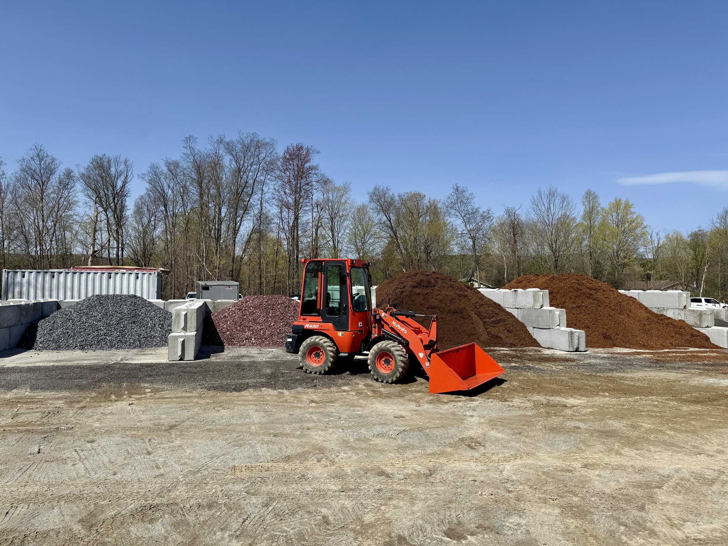 Construction site with a small orange bulldozer in front of piles of gravel, dirt, and concrete blocks, with trees and a blue sky in the background.