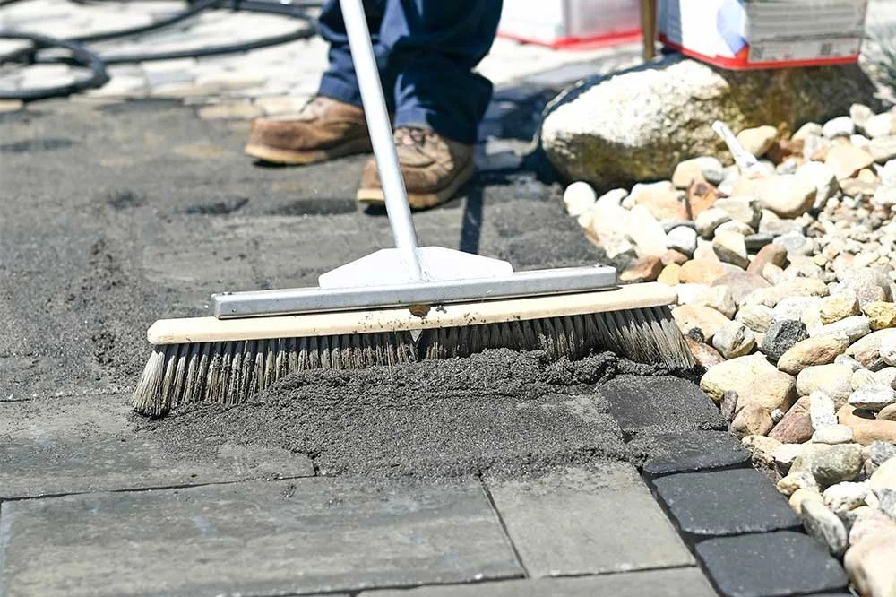 A person using a push broom to sweep fresh asphalt during a paving project. Rocks and paving stones are visible on the side.