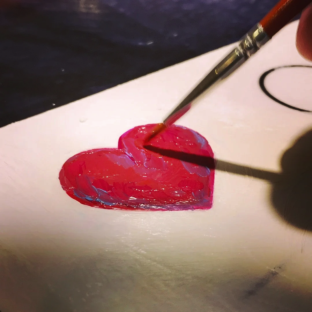 A person painting a red heart with a paintbrush on a white surface.