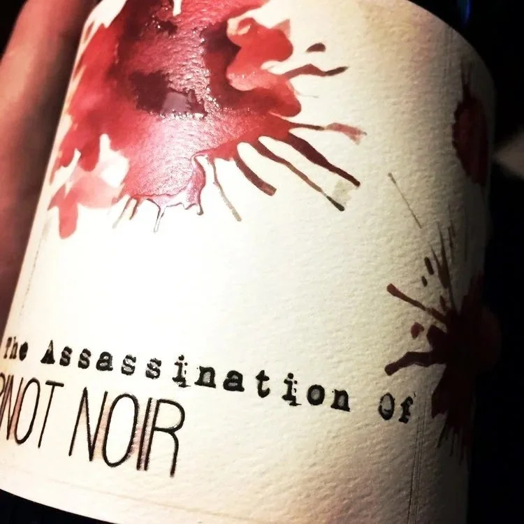 Close-up of a wine label with watercolor flower artwork, the text 'the assassination of' and 'NOT NOR' visible.