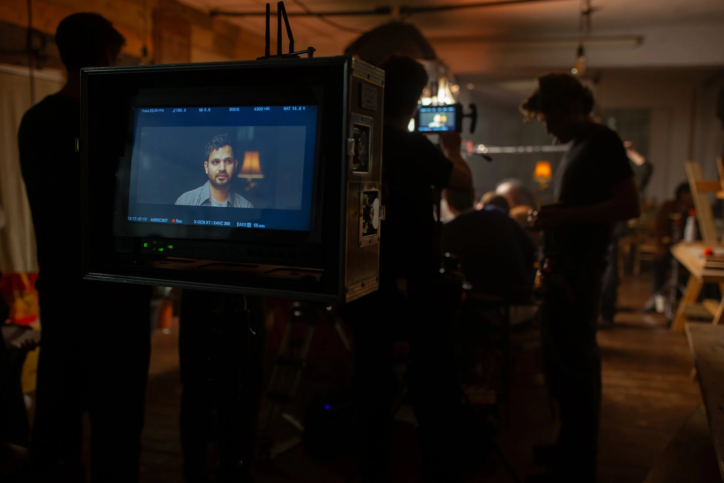 On set of a film or video production, a camera focusing on a male actor with dark hair and a beard, with crew members working in a dimly lit, rustic room with wooden walls and warm lighting.