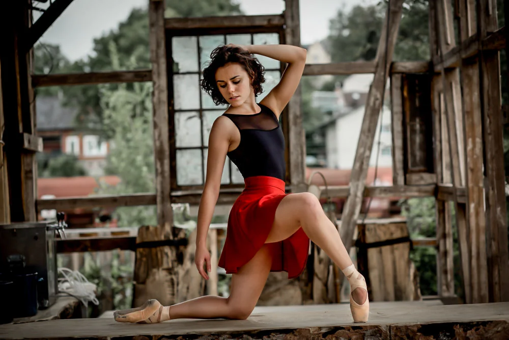 A young woman in ballet attire posing on a wooden stage in an unfinished, rustic wooden building with a window and outdoor trees visible in the background.