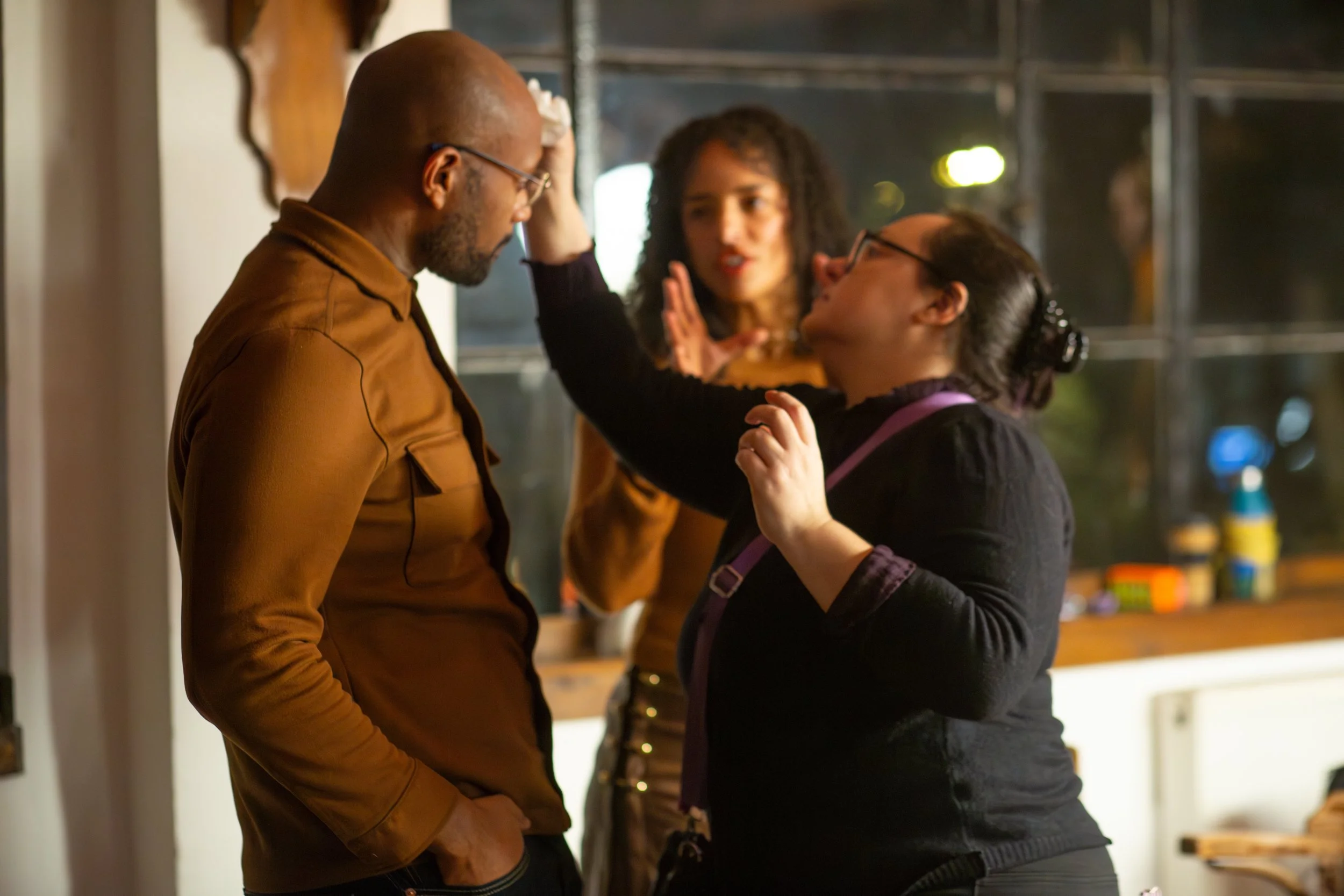Three people having a serious conversation indoors at night, with the woman in glasses gesturing with her hand.