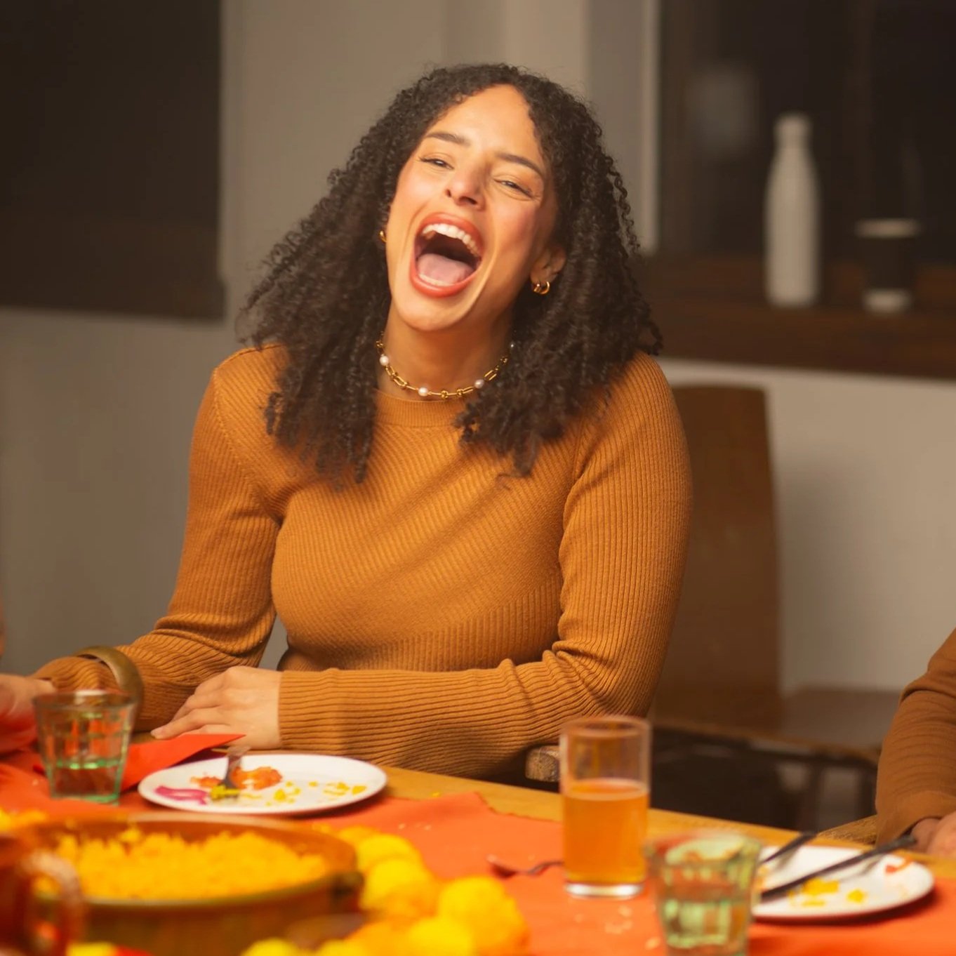 A woman with curly hair, wearing a mustard-colored sweater and jewelry, laughing during a social gathering.