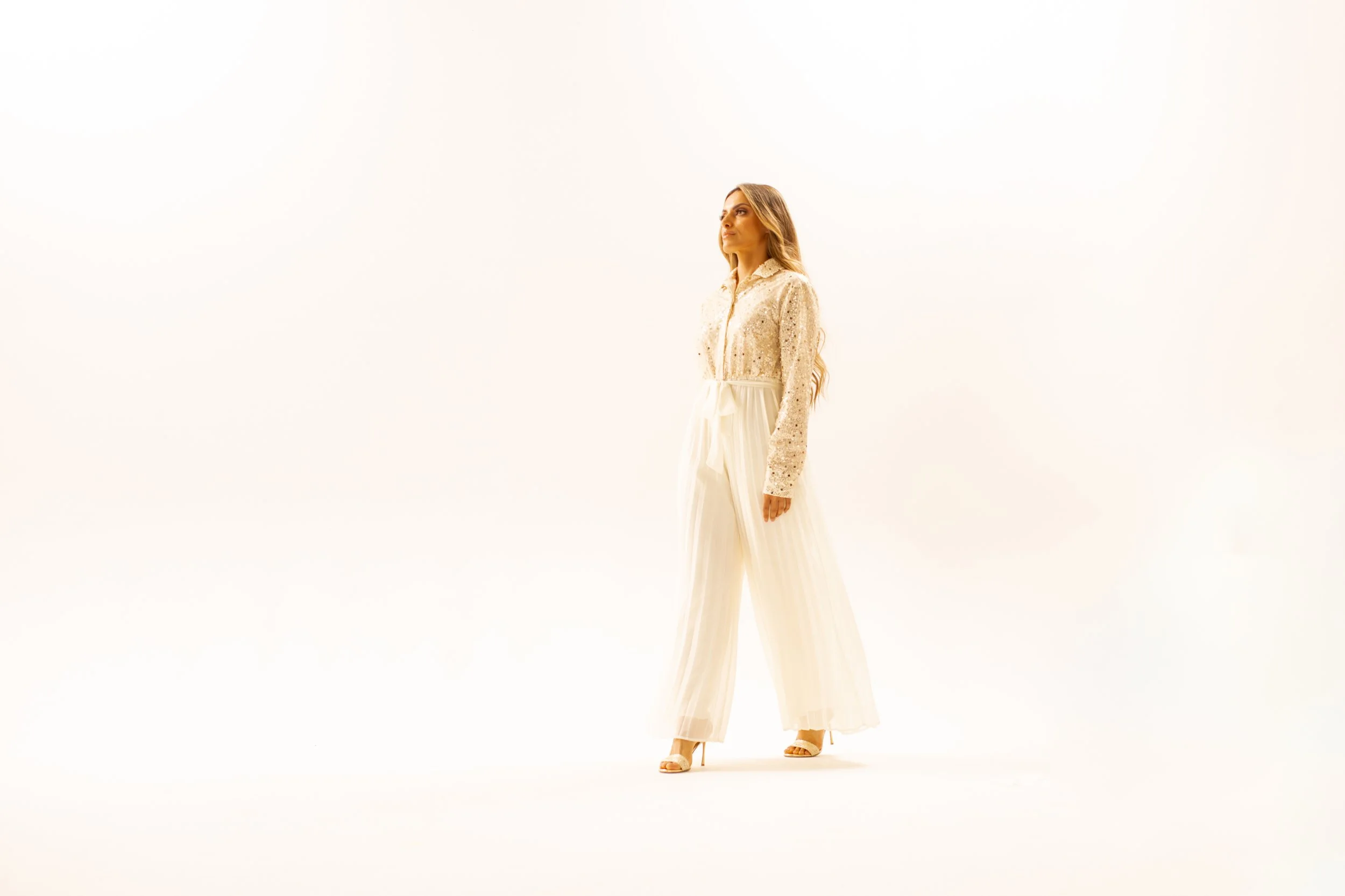 A woman with long, wavy hair standing against a plain white background. She is wearing a cream-colored, long-sleeved, sequined blouse and wide-leg, high-waisted pants, with beige heels.