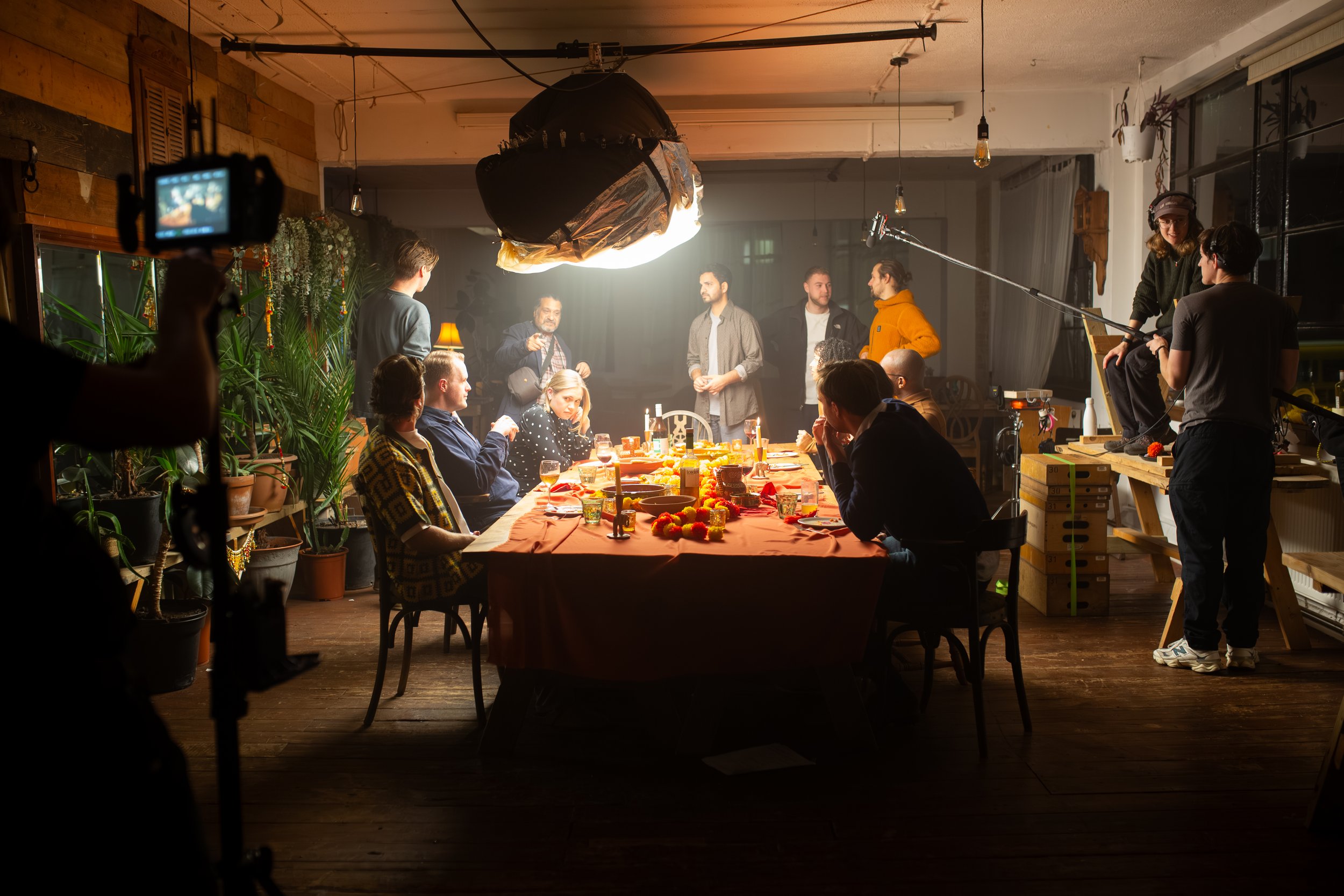 Behind-the-scenes of a video shoot or film production in a cozy indoor setting with warm lighting and a long dinner table decorated with flowers and food, crew members and actors preparing and filming around the table.