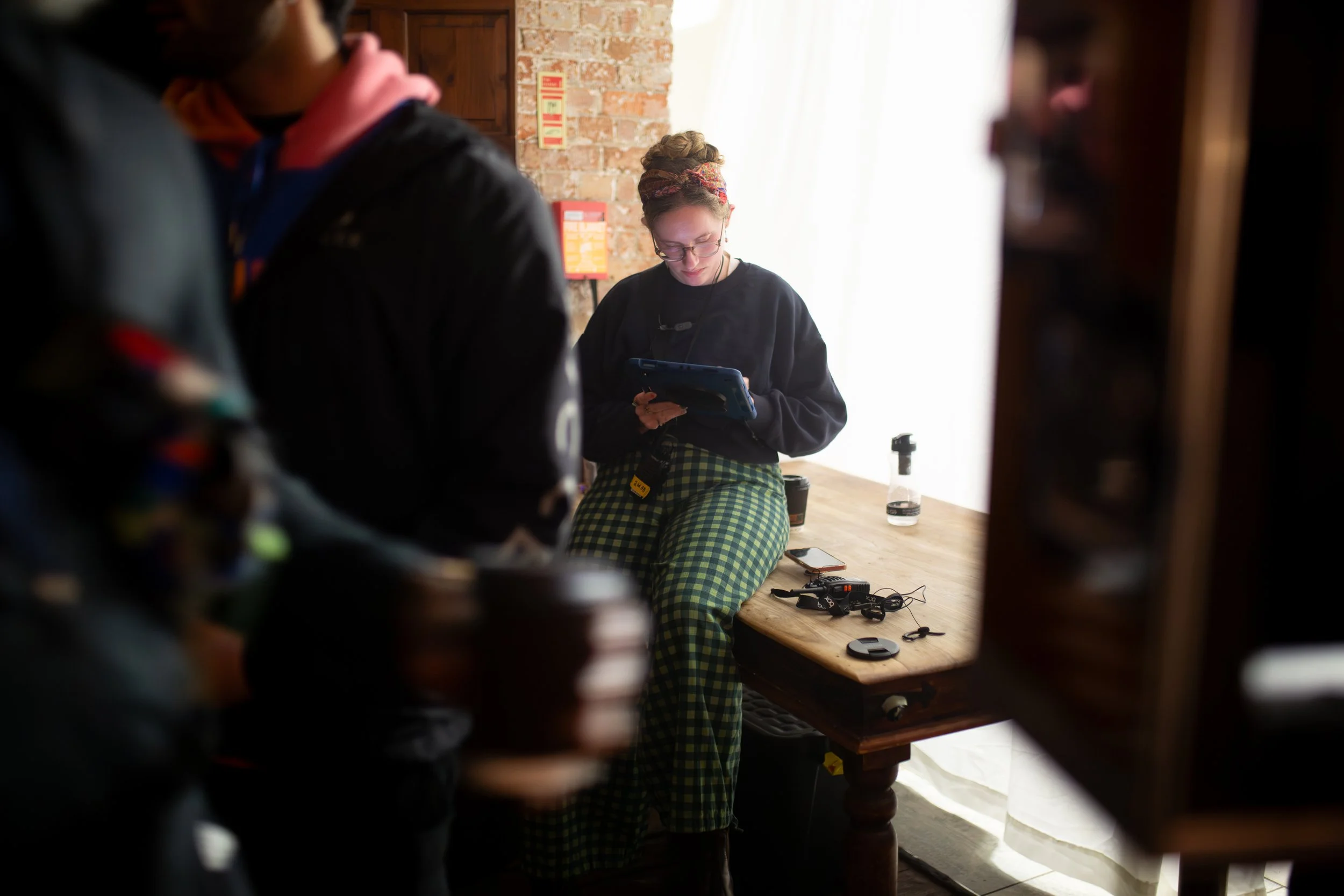 A young woman sitting at a wooden table, looking at her phone, wearing glasses and colorful checkered pants, with a brick wall and natural light behind her. Others are partially visible in the foreground, out of focus.