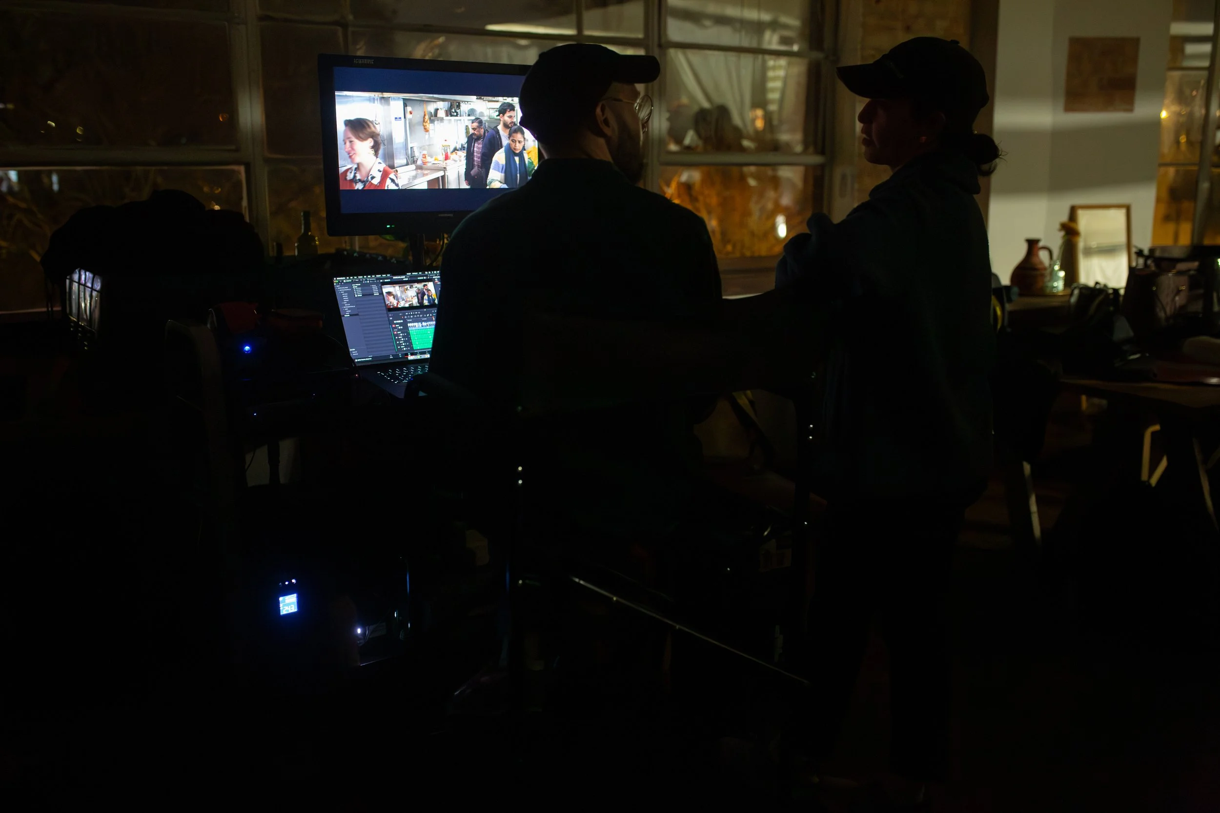 Two people working on video editing or production in a dimly lit room with a computer displaying a video editing software. A monitor shows people in a kitchen or restaurant setting.
