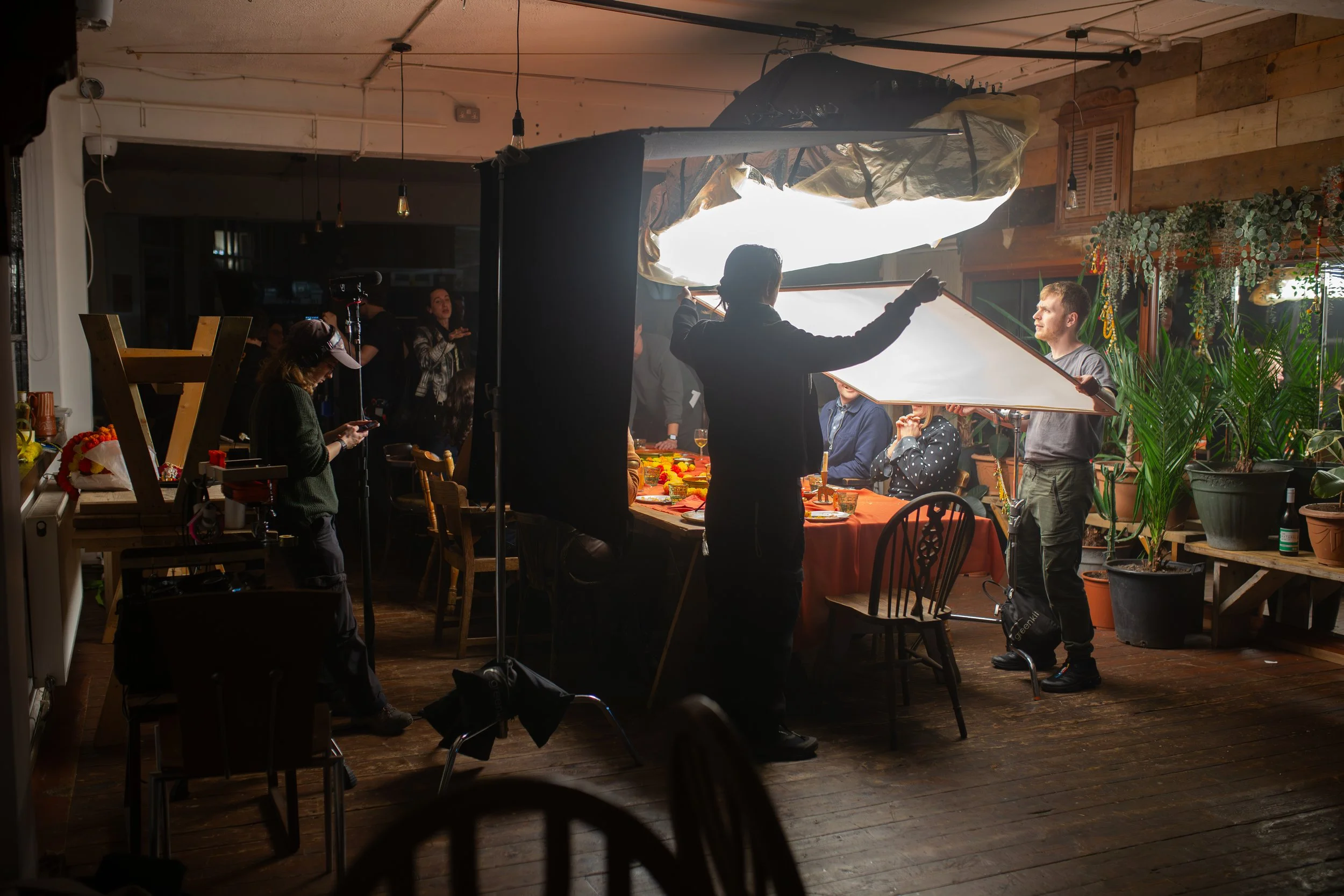 A filming or photography setup indoors with a large light diffuser, crew members adjusting equipment, a person in a gray shirt sitting at a table, and various plants and decorative items in the background.