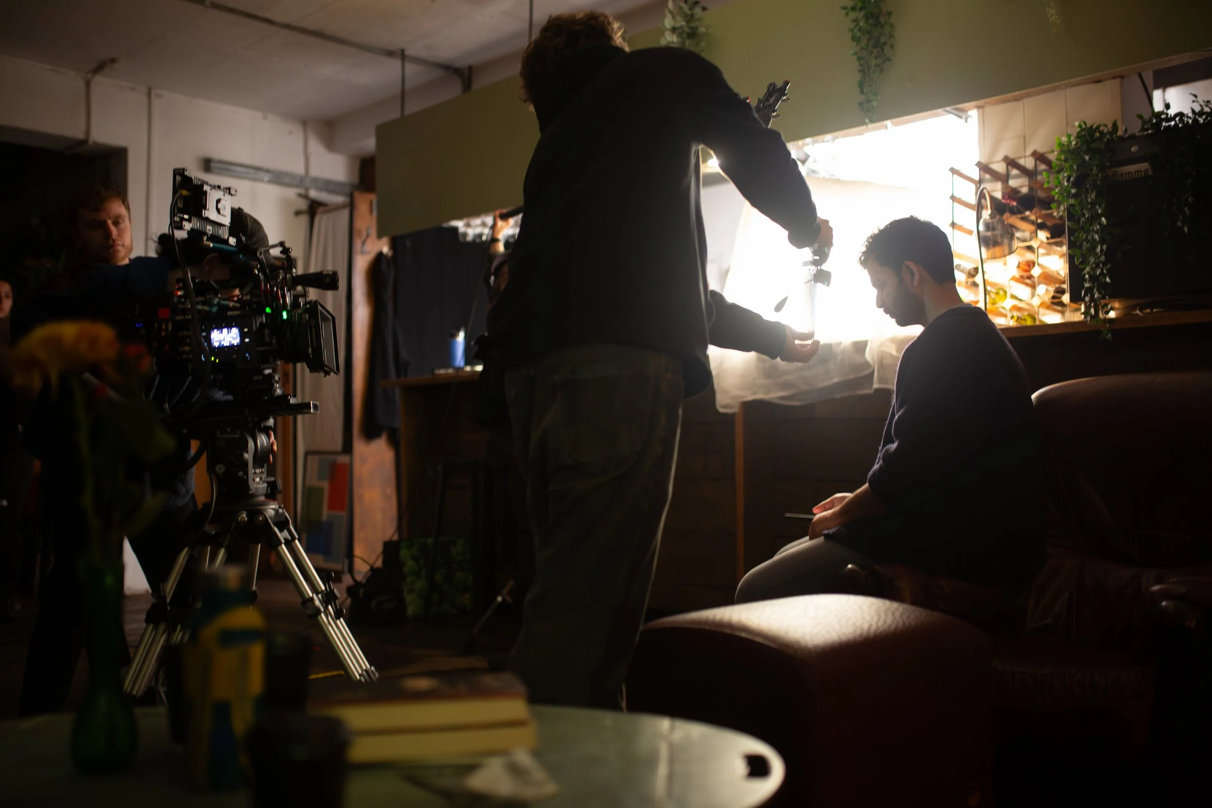 Behind-the-scenes shot of a film or photo shoot in a cozy, warm-lit indoor space. A man is seated on a chair near a window, with the silhouette of another person adjusting equipment or preparing for a shot. The room features wooden shelves with plant