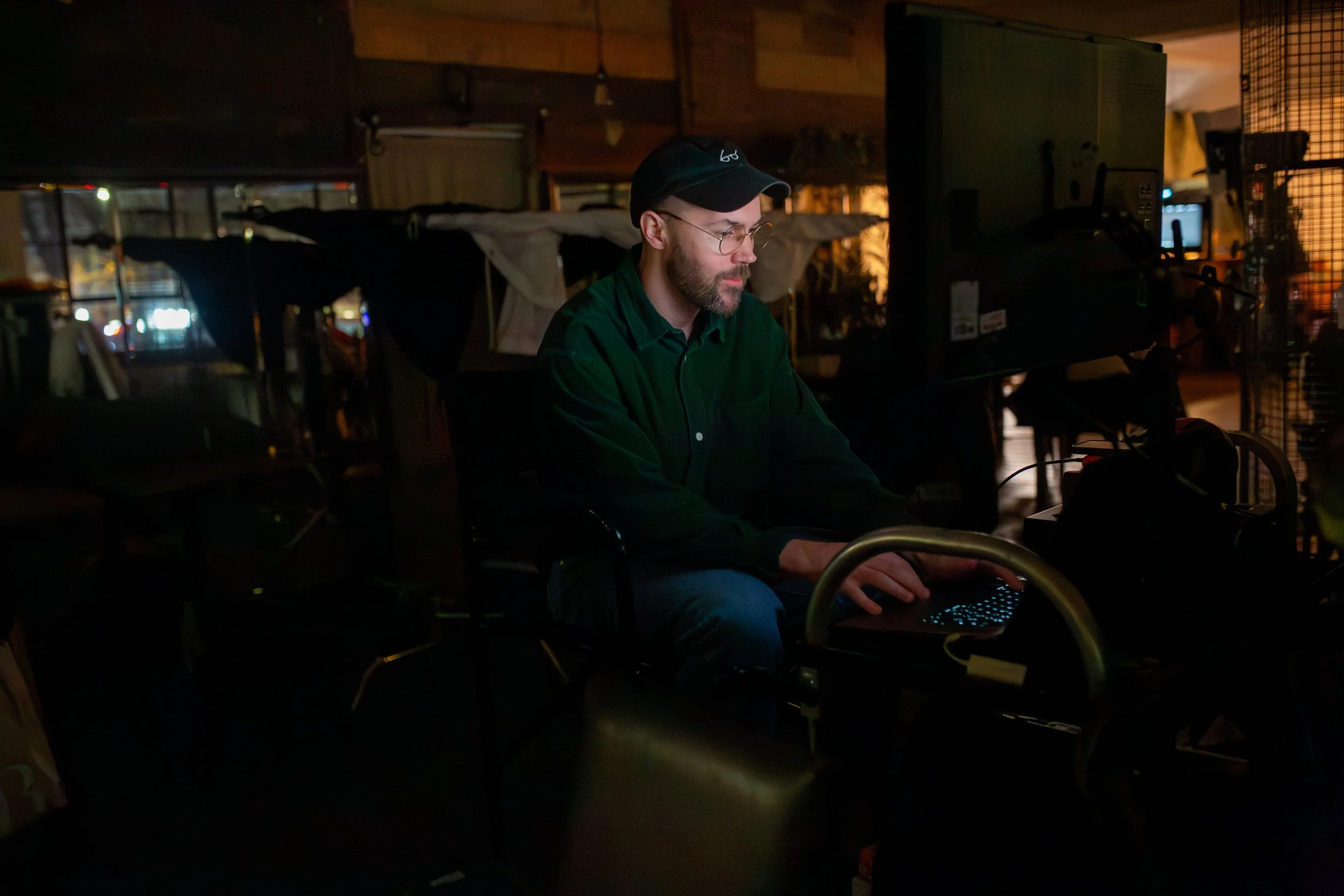 A man with glasses, a beard, wearing a black cap and green shirt, working on a laptop in a dimly lit indoor space with wooden walls and various electronic equipment around him.