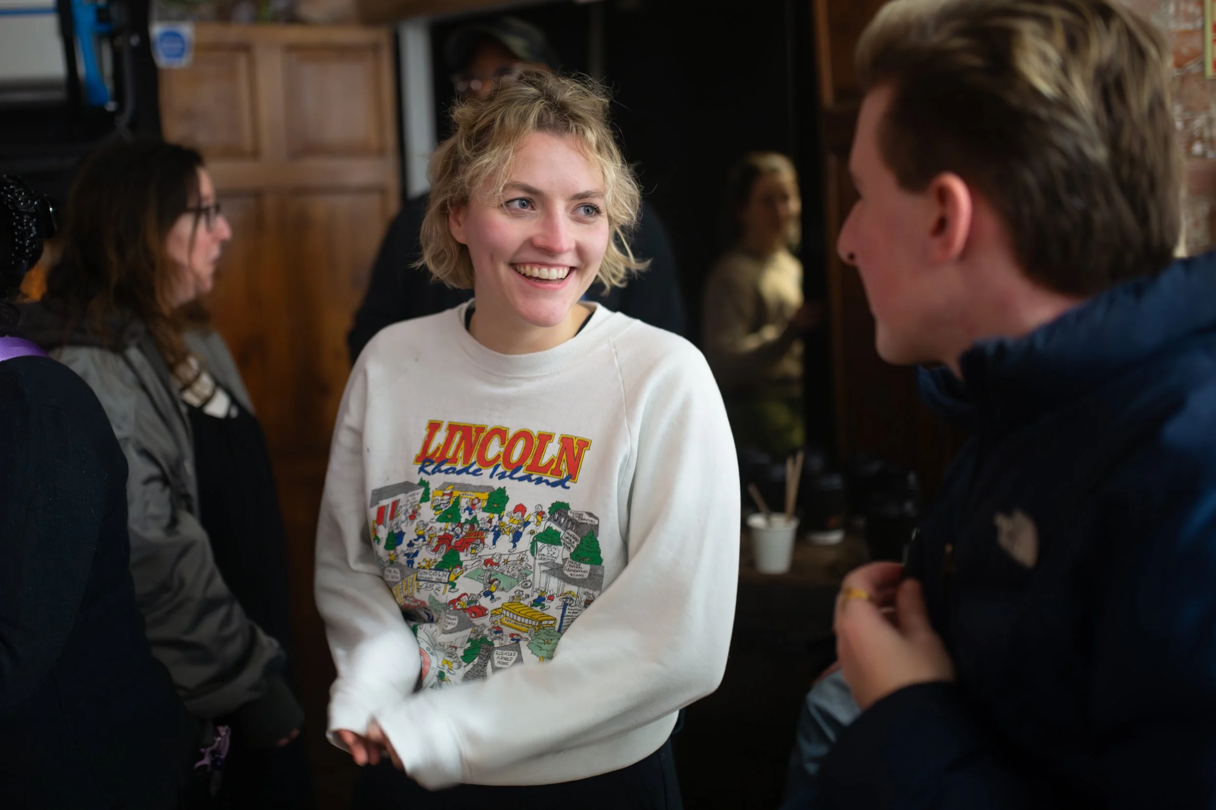A woman with short blond hair smiling and talking to a person with short brown hair in a crowded indoor setting, possibly a social event or gathering.