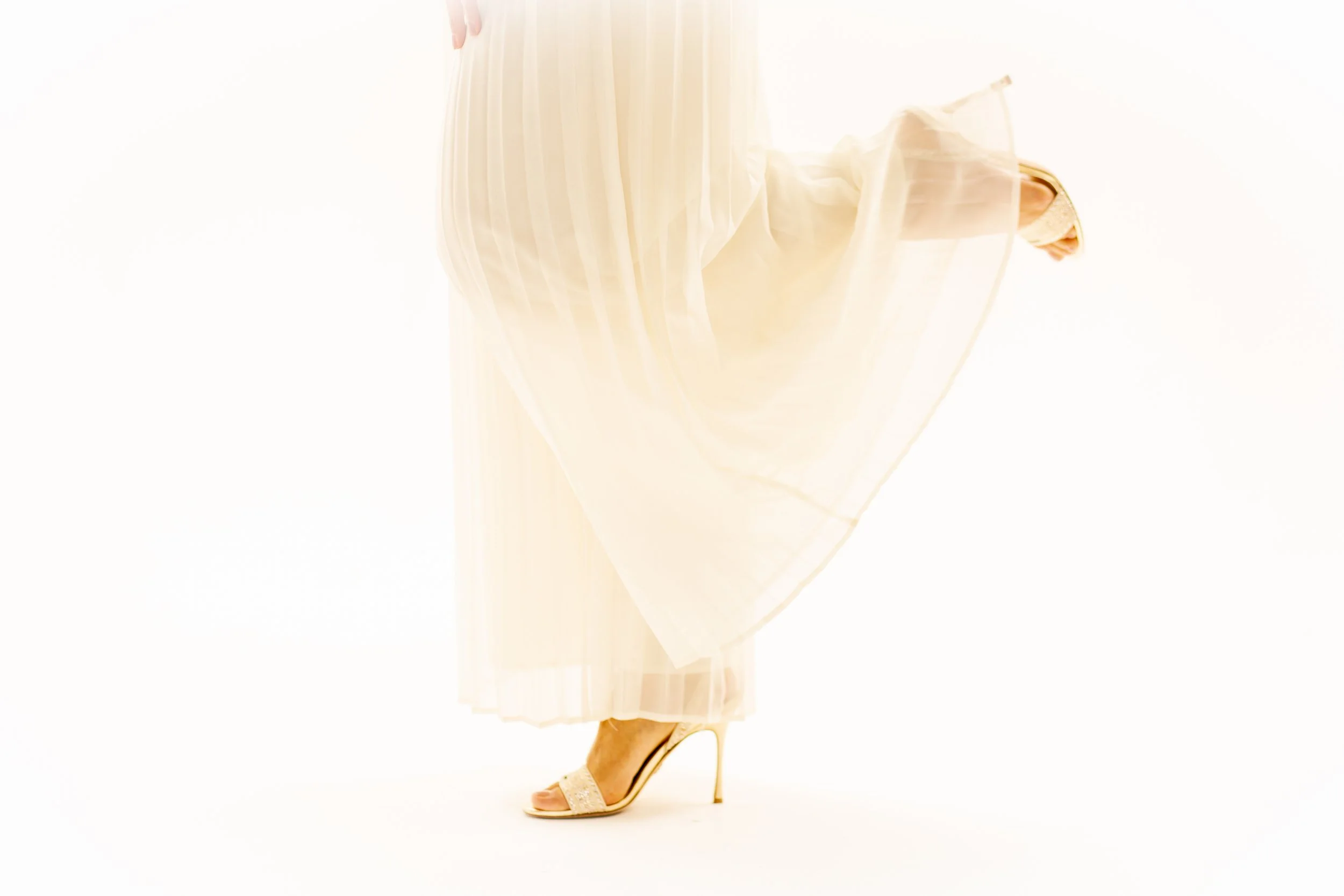 Close-up of a woman wearing a white dress and high-heeled shoes, showing only her lower body and legs against a white background.