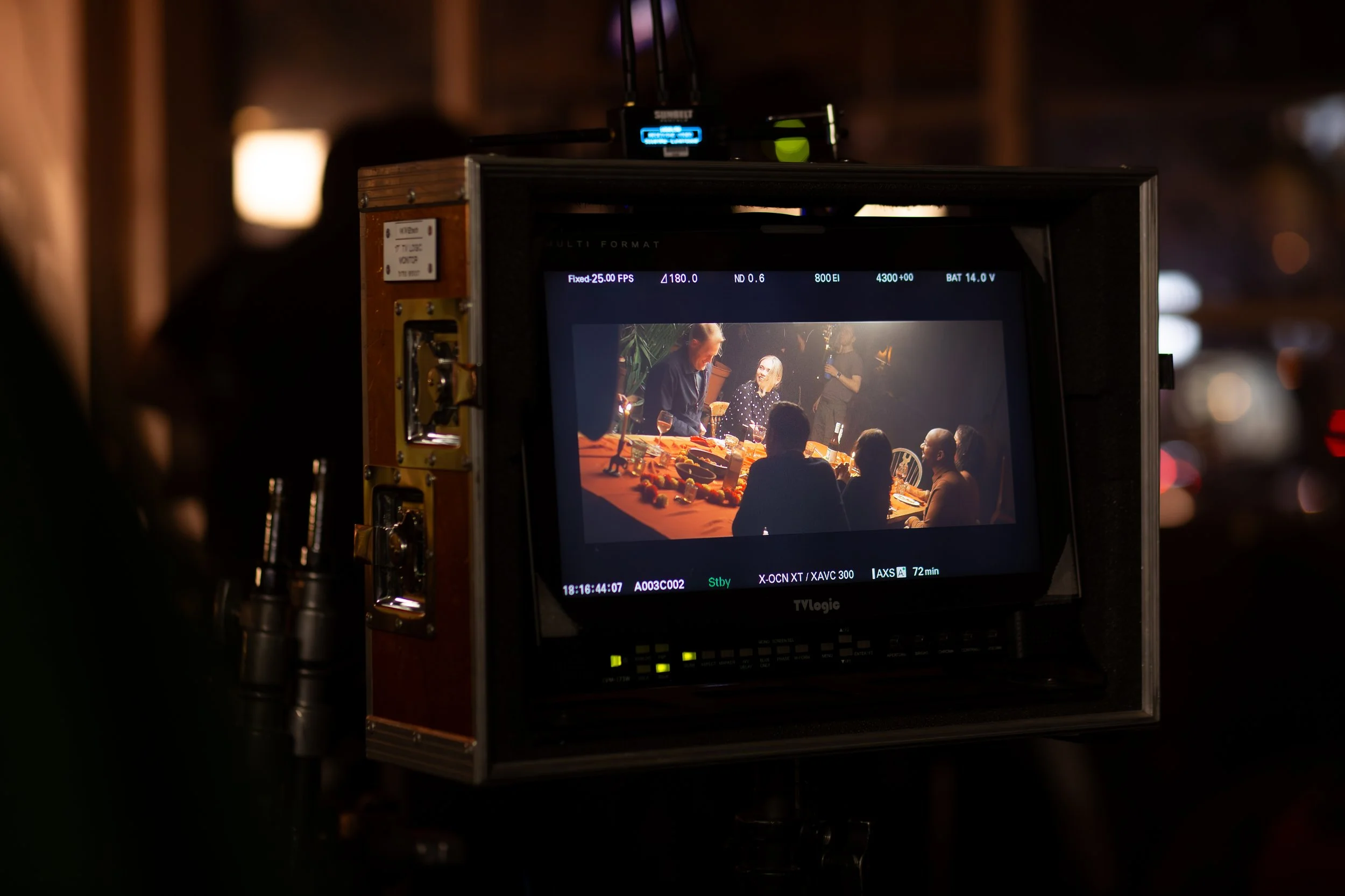 View of a camera monitor capturing a scene of several people gathered around a table, with one person standing and speaking, in a dimly lit indoor setting.